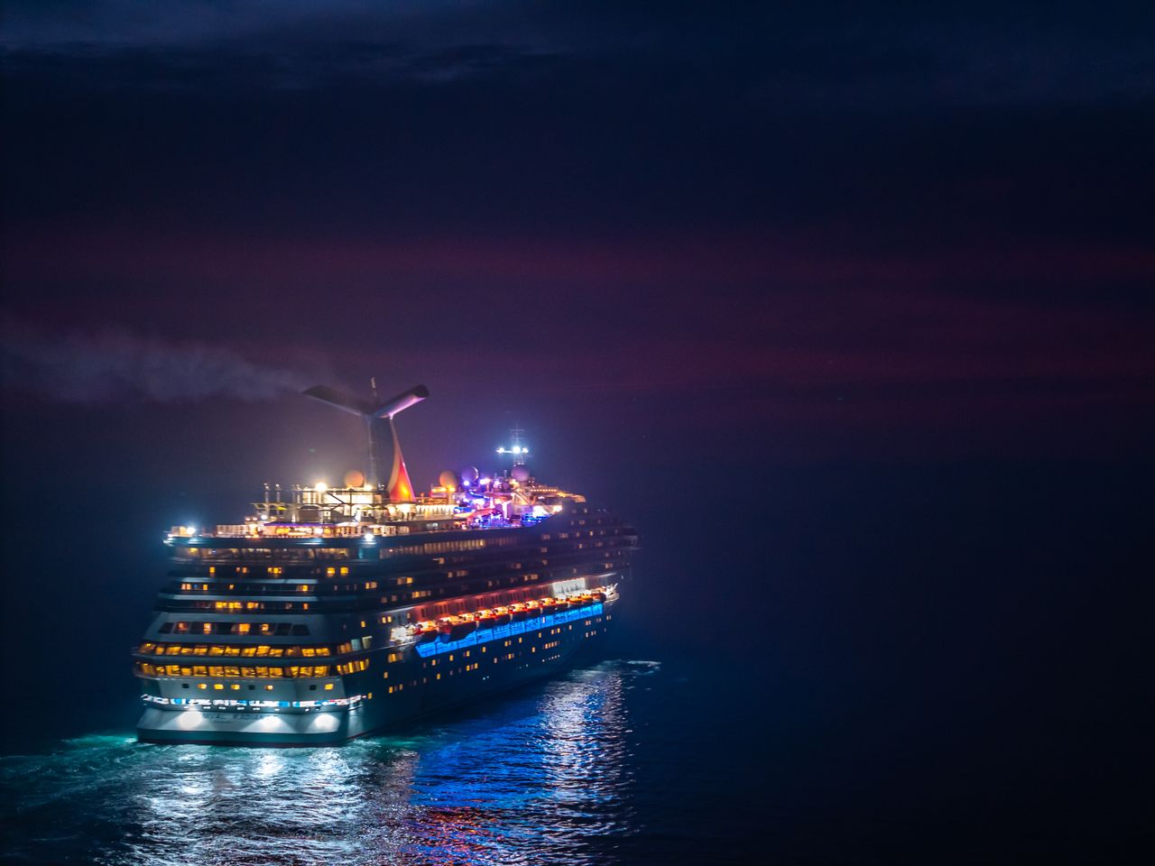 A brightly lit cruise ship sails through the ocean under a vivid night sky.
