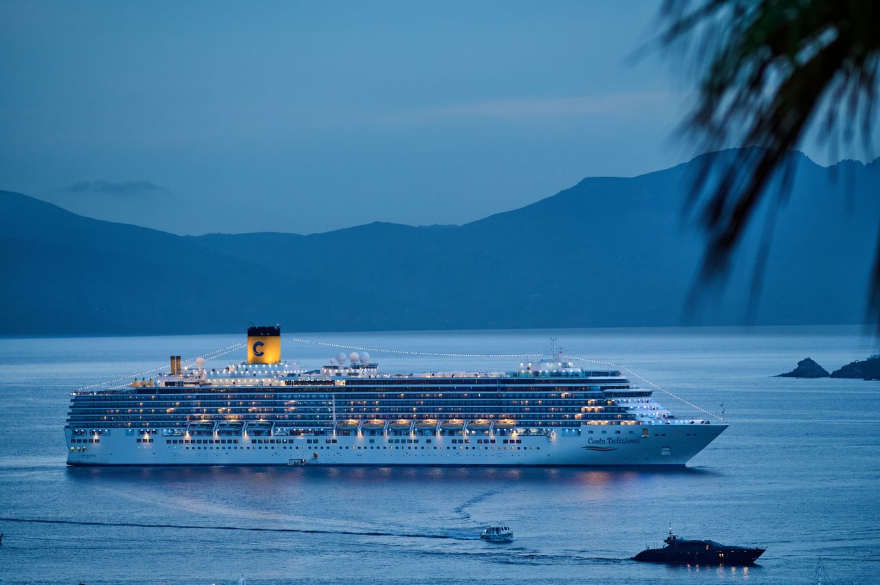 A beautifully illuminated cruise ship sails across the calm sea at dusk, framed by mountains.